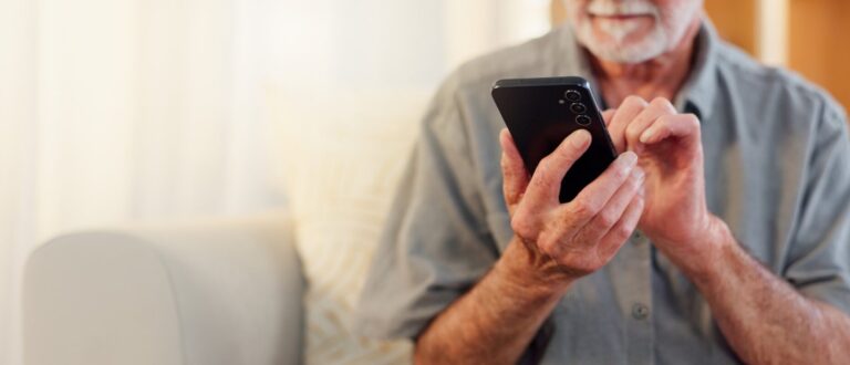 Oudere man gebruikt smartphone in zijn woonkamer – symbool voor digitale zelfstandigheid en gebruiksvriendelijke technologie voor senioren
