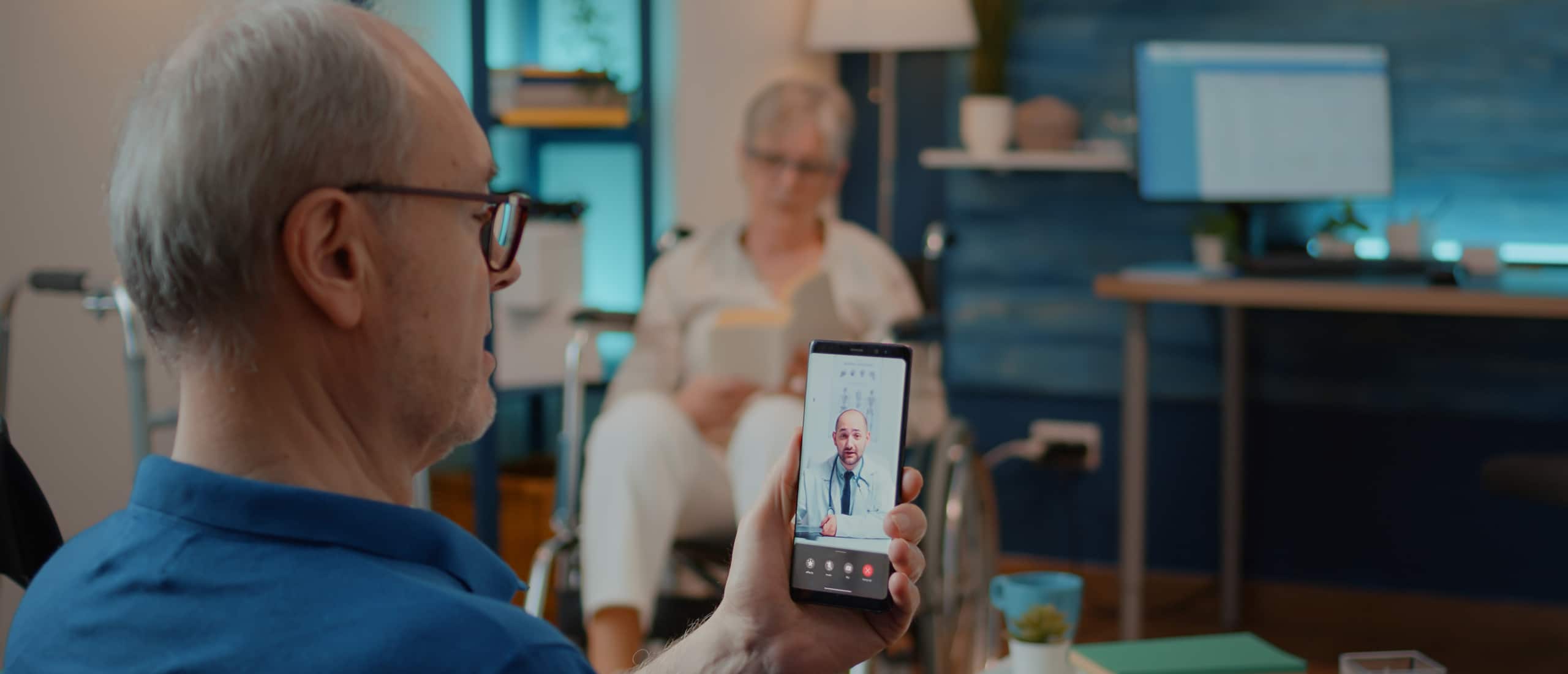 Een oudere man gebruikt zijn smartphone om te videobellen met een arts, terwijl hij thuis in de woonkamer zit – zorgtechnologie in actie.